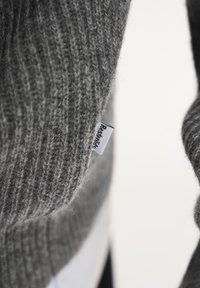 Grå strikket sweater med ribbet tekstur. Har et lille stofmærke syet på siden, der viser brandnavnet i sort tekst.