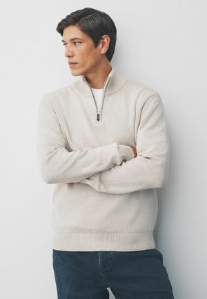 Young man with dark hair wearing a beige half-zip sweater and dark jeans stands with arms crossed, looking to his left against a plain background.