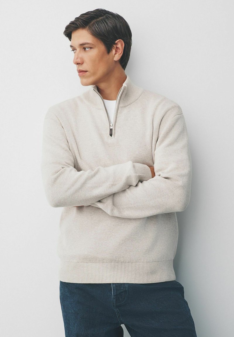 Jonge man met donker haar in een beige half-zip trui en donkere spijkerbroek staat met gekruiste armen, kijkend naar links tegen een effen achtergrond.