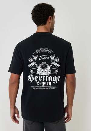 Mand iført sort T-shirt med hvid vintage motorcykelgrafik og teksten "Legendary Bike Co. Born to Inspire Heritage Legacy" på bagsiden.