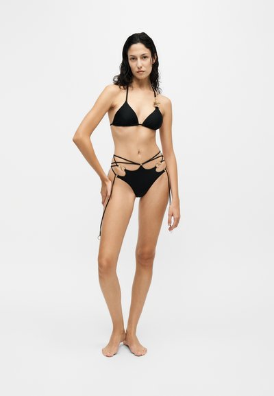 Femme aux longs cheveux foncés portant un bikini noir à bretelles avec des détails en anneau doré, debout pieds nus devant un fond clair uni.