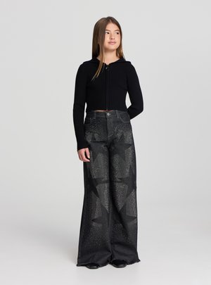 Czarna prążkowana krótka bluza z kapturem i zamkiem z przodu, zestawiona z czarnymi szerokimi spodniami z wzorem gwiazdek i ćwiekami.