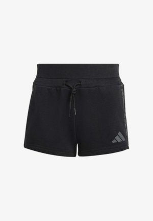 Niña vistiendo ropa deportiva negra de Adidas, pantalones cortos negros, calcetines a rayas y zapatillas, sosteniendo una chaqueta sobre el hombro y sonriendo.