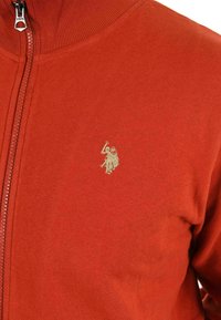 Roter Reißverschluss-Pullover aus weichem Stoff mit geripptem Kragen. Verfügt über ein kleines goldenes Logo auf der linken Brustseite. Glatte Textur.