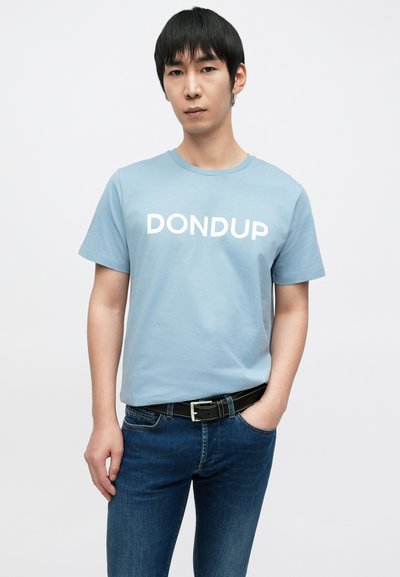 Ανοιχτό μπλε μπλουζάκι με τη λέξη "DONDUP" σε έντονα λευκά γράμματα, συνδυασμένο με σκούρο μπλε τζιν παντελόνι και μαύρη ζώνη με ασημένιες λεπτομέρειες.