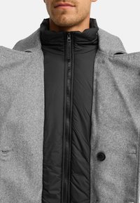 Abrigo de lana gris con una capa interna negra con cremallera. Presenta un cuello alto, detalles de botones y un acabado texturizado en el material exterior.