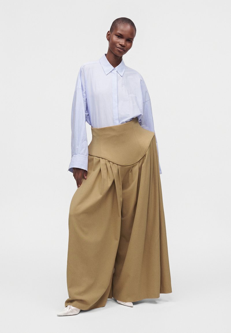 Mannequin portant une chemise oversize bleu clair rentrée dans un pantalon beige taille haute à jambes larges avec plis et chaussures blanches pointues sur fond blanc.