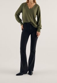 Suéter de punto texturizado en verde oliva con cuello en V y puños de manga fruncidos, combinado con jeans negros de campana y botas de tobillo puntiagudas en negro.