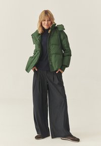 Jachetă pufos verde cu glugă, închidere cu fermoar și model matlasat; asortată cu pantaloni largi cu dungi și pantofi maronii.