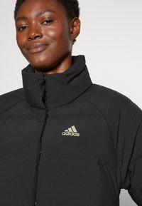 Czarna puchowa kurtka z wysokim kołnierzem, z srebrnym logo Adidas na piersi. Gładka tekstura i minimalistyczny design.