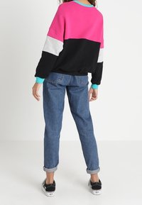 Sweat-shirt coloré avec des panneaux roses, gris et noirs, un col turquoise, associé à un jean bleu retroussé et des baskets noires.