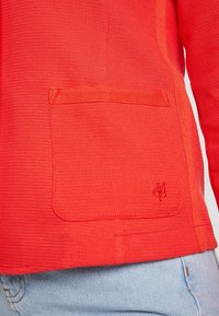 Czerwony sweter o fakturowanej powierzchni z kieszenią z przodu, z subtelnymi detalami ściegów i małym logo na dole. Luźny krój, zestawiony z jasnymi dżinsami.