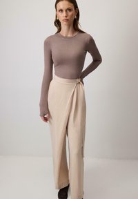 Woman wearing taupe long-sleeve fitted top and beige wrap-style wide-leg pants with buckle detail, standing against a plain light background.