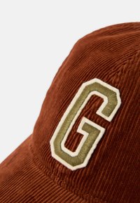 Braune Corduroy-Mütze mit einem grünen, in Weiß umrandeten gestickten Buchstaben "G", die eine strukturierte Form und eine texturierte Oberfläche aufweist.