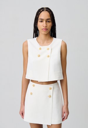 Top corto bianco con bottoni floreali dorati e due tasche frontali, abbinato a una minigonna coordinata con bottoni simili e un design avvolgente.