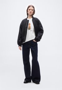 Γυναίκα με μαύρο bomber μπουφάν, λευκό μπλουζάκι με γραφικό σκύλου, σκούρο φαρδύ τζιν και μαύρα παπούτσια, που στέκεται μπροστά σε λευκό φόντο.