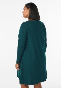 Žena s rovnými čiernymi vlasmi, oblečená v dlhých rukávoch, šatách do kolien v tmavo tyrkysovej farbe, stojí na boku pred bielym pozadím.