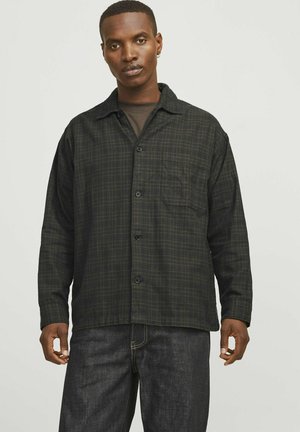 Camisa de franela a cuadros en verde oscuro y negro, de manga larga, con botones en la parte delantera, un bolsillo en el pecho y llevada sobre una camiseta oscura, combinada con jeans oscuros.