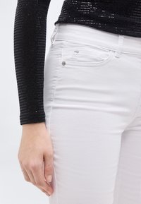Gros plan d'une personne portant un jean blanc et un haut à manches longues noir avec des détails en sequins, main détendue le long du corps.