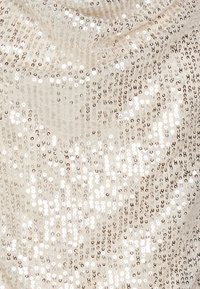 Beige tkanina pokrytá lesklými flitry, odrážející světlo s hladkou strukturou. Povrch zobrazuje hustý vzor kulatých ozdob.