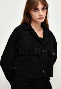 Schwarze Tweedjacke mit strukturiertem Finish, ausgestattet mit einem Kragen, Fronttaschen und goldfarbener Hardware. In einem kurzen Design gestaltet.