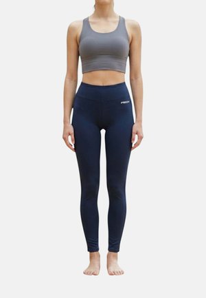 Donna che indossa un reggiseno sportivo grigio e leggings blu navy a vita alta, in piedi a piedi nudi su sfondo bianco.