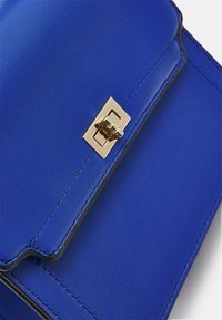 Blå läderhandväska med scalloped flap-kant och rektangulär guldmetallspänne.