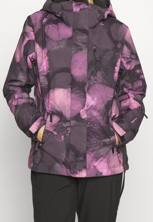 Persoon die een paarse en zwarte patroon winterjas met capuchon draagt, met handen in de zijzakken en een zwarte broek.