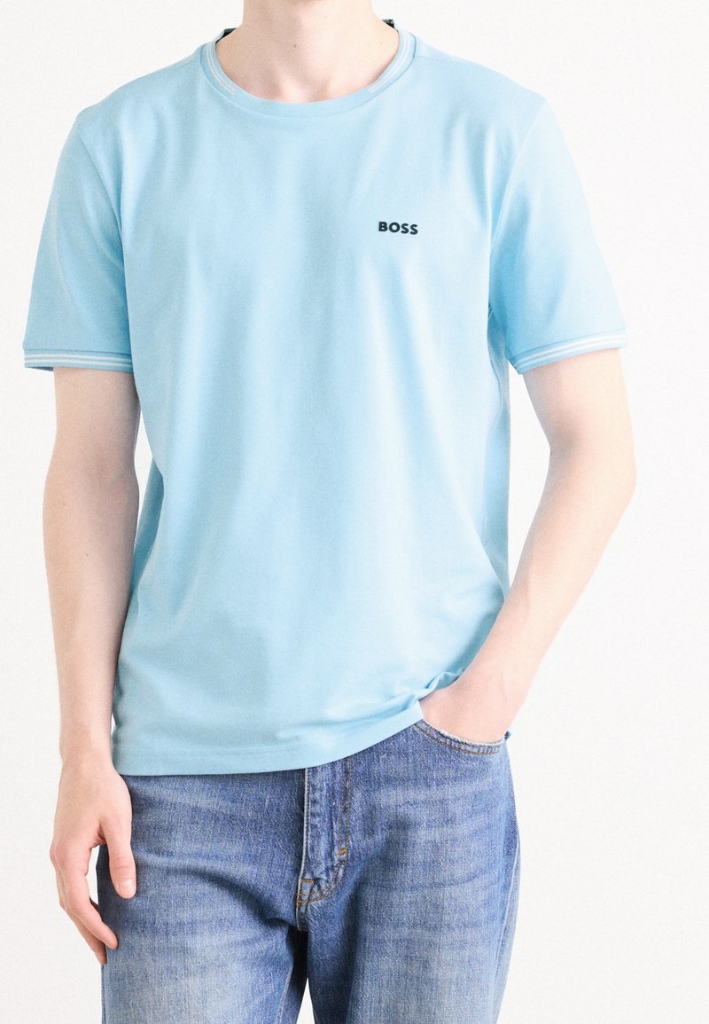Personne portant un T-shirt à manches courtes bleu clair avec le texte « BOSS » sur la poitrine et un jean bleu, debout avec une main dans la poche.