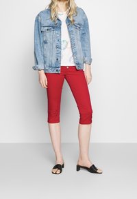 Vaaleansininen farkkutakki, jossa kaksi rintataskua. Valkoinen t-paita, jossa monivärisiä pyöreitä kuvioita. Punaiset caprihousut. Mustat liukusandaalit.