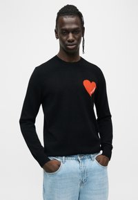 Pull noir en tissu tricoté avec un motif de cœur rouge sur la poitrine gauche. Le modèle le porte avec un jean en denim bleu clair.