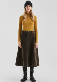Vrouw met een zwarte gebreide muts, mosterdkleurige coltrui, donkerbruine ribfluwelen midi-jurk en zwarte kniehoge laarzen op een effen achtergrond.