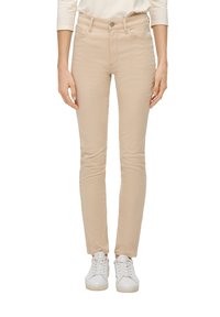 Jeans skinny beige fabricados en tejido elástico, con cintura de altura media, bolsillos delanteros y cierre de botón, combinados con zapatillas blancas.
