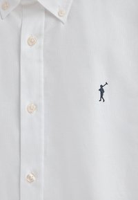 Camicia bianca button-down con colletto classico, realizzata in tessuto testurizzato. Presenta bottoni chiari e un piccolo logo ricamato blu navy raffigurante una figura che tiene un bastone.