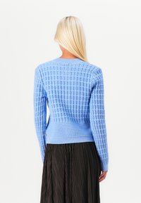 Šviesiai mėlynas megztinis su tekstūruotu raštu, apvaliu iškirpimu ir raukšlėtais rankogaliais, derinamas su plisuota juoda sijonu.