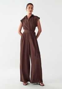 Bruine, mouwloze jumpsuit met een kraag en een ceintuur in de taille, en een ontwerp met wijde pijpen. Gemaakt van een gladde stof met twee zijzakken.