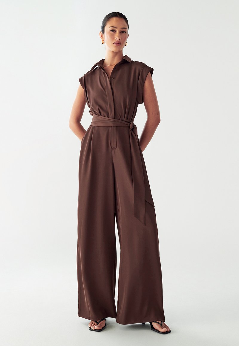 Brun, ærmeløs jumpsuit med kraveudskæring, belted talje og bred ben-design. Fremstillet af glat stof med to sidelommer.