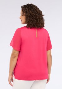 Krøllet hår kvinde iført lys pink kortærmet bluse med nøglehul på ryggen, stående sidelæns foran en ensfarvet baggrund.