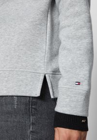Manga de sudadera gris claro con puño de canalé azul marino, pequeño logo de Tommy Hilfiger en el puño y la manga, usado sobre unos jeans de mezclilla negros.