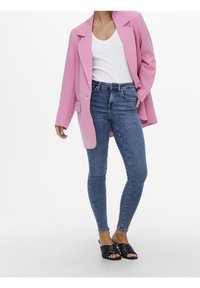 Woman wearing a pink blazer, white tank top, blue skinny jeans, and black heeled sandals standing with one hand in pocket.