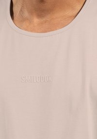 Světle béžové tričko s širokým výstřihem a měkkou texturou. Na hrudi je reliéfní logo značky "SMILODOX".