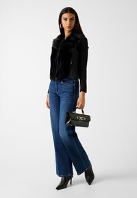 Czarna kamizelka z futerka ekologicznego z teksturowanym tyłem, zestawiona z niebieskimi spodniami o szerokich nogawkach. Modelka trzyma czarną torebkę ze złotymi zdobieniami i nosi czarne szpilki.