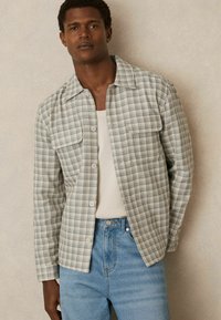 Cămașă cu carouri ușoare în verde și crem, cu două buzunare la piept și o croială relaxată, asortată cu blugi din denim albastru deschis.