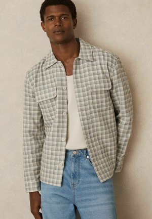 Cămașă cu carouri ușoare în verde și crem, cu două buzunare la piept și o croială relaxată, asortată cu blugi din denim albastru deschis.