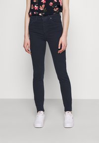 Jeans skinny de mezclilla oscura con talle alto, que presentan un diseño clásico de cinco bolsillos y un ligero desvanecimiento. Combinados con una blusa floral y zapatillas blancas.