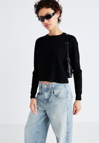 Zwarte cropped sweatshirt met lange mouwen, gecombineerd met lichtblauwe high-waisted jeans. Accessoires omvatten een kleine zwarte handtas en een zonnebril.