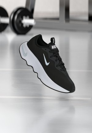 Czarno-biały sneaker Nike z zaokrągloną podeszwą unoszący się nad błyszczącą podłogą, na tle rozmytych ciężarków siłowni.