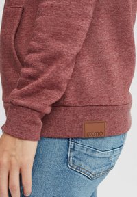 Haut à capuche bordeaux, fabriqué en tissu texturé, avec une poche avant, des poignets côtelés et un patch logo en cuir marron près de l'ourlet.