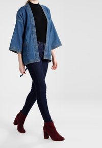 Chaqueta kimono de mezclilla con mangas anchas y dos bolsillos frontal, llevada sobre una blusa negra, combinada con jeans ajustados azul marino y botas de tacón burdeos.