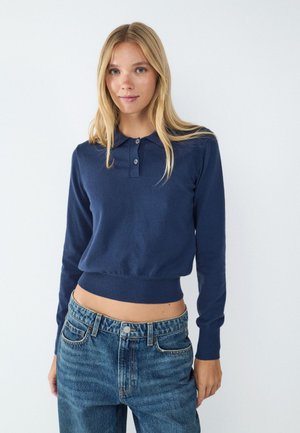 Jeune femme aux longs cheveux blonds porte un pull polo bleu marine à manches longues et un jean bleu taille haute sur un fond blanc uni.